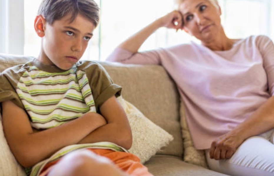 Teenage boy has arms folded and looks unhappy and defiantly away as nearby parent appears to be talking to him