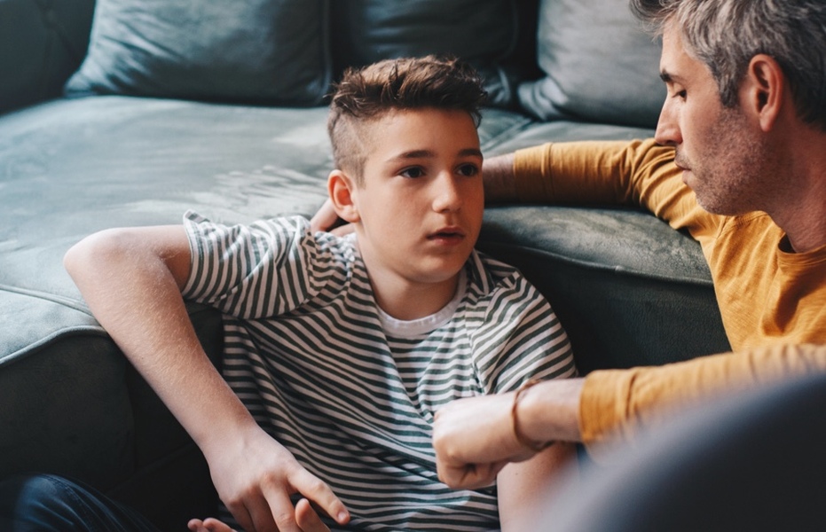 Teenage boy sits on the floor in front of couch with parent sitting beside him, looking concerned