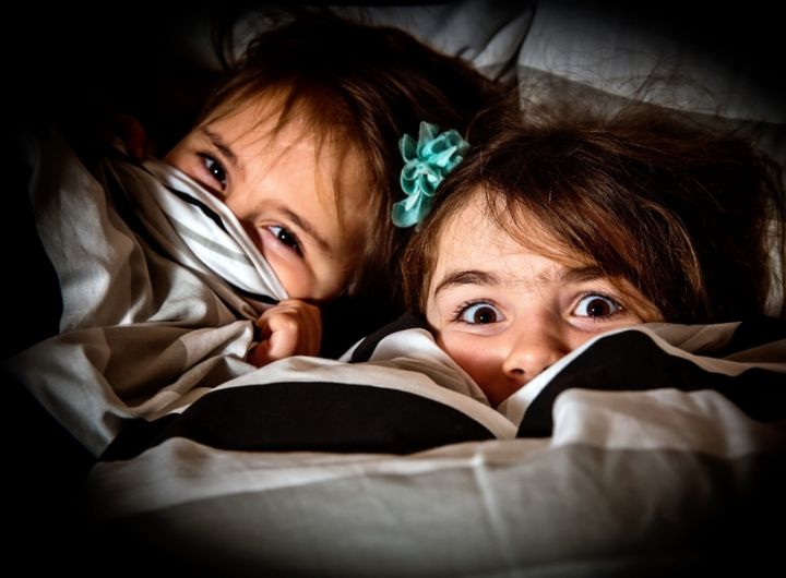 girls hiding under the bedcovers - one scared, one smiling