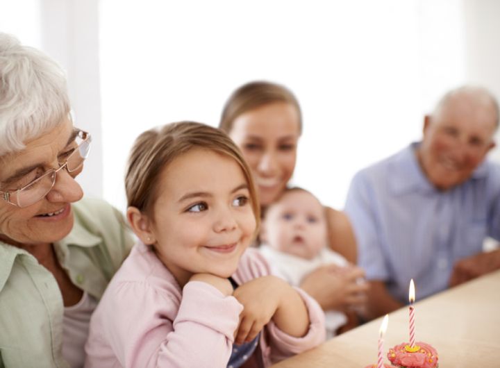 granddaughter ready to blow out cake candle sits on grandmother's lap, other family look on